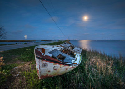 An abandoned boat lays washed up on the island road, with the lights of Point Aux Chene on the horizon. The road that links the islanders to nearby towns now floods regularly.